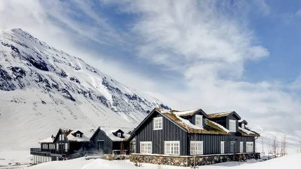Dark timber lodge buildings with snow-covered roofs at the foot of a mountain.