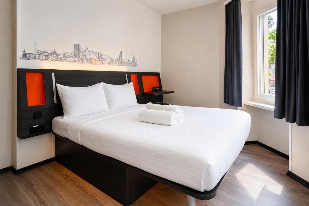 A narrow hotel room with a bed against a dark headboard and a window to one side.
