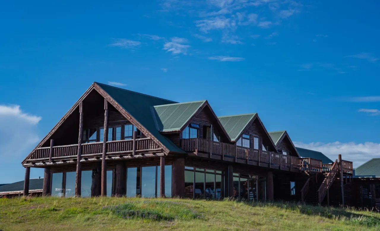 A dark log lodge with green gabled roofs and a glass ground floor on a grassy slope.