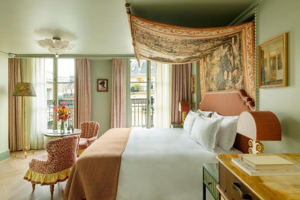 A hotel bedroom with pale walls, a tapestry canopy over the bed and a view of city rooftops.